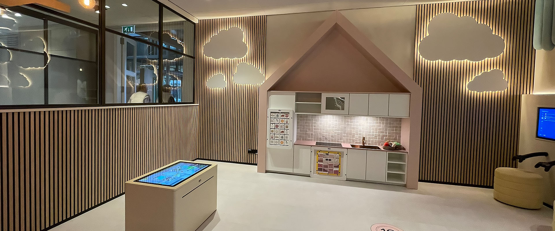 Een sfeervol ingerichte kinderhoek met een speelkeuken in een huisvormig thema, interactieve vloer met hinkelcirkels en een digitale speeltafel. De wand is versierd met houten latten en wolkvormige lichtpanelen. De ruimte is warm verlicht en bevat speelse elementen zoals een houten boomstructuur aan het plafond en retro lampen.