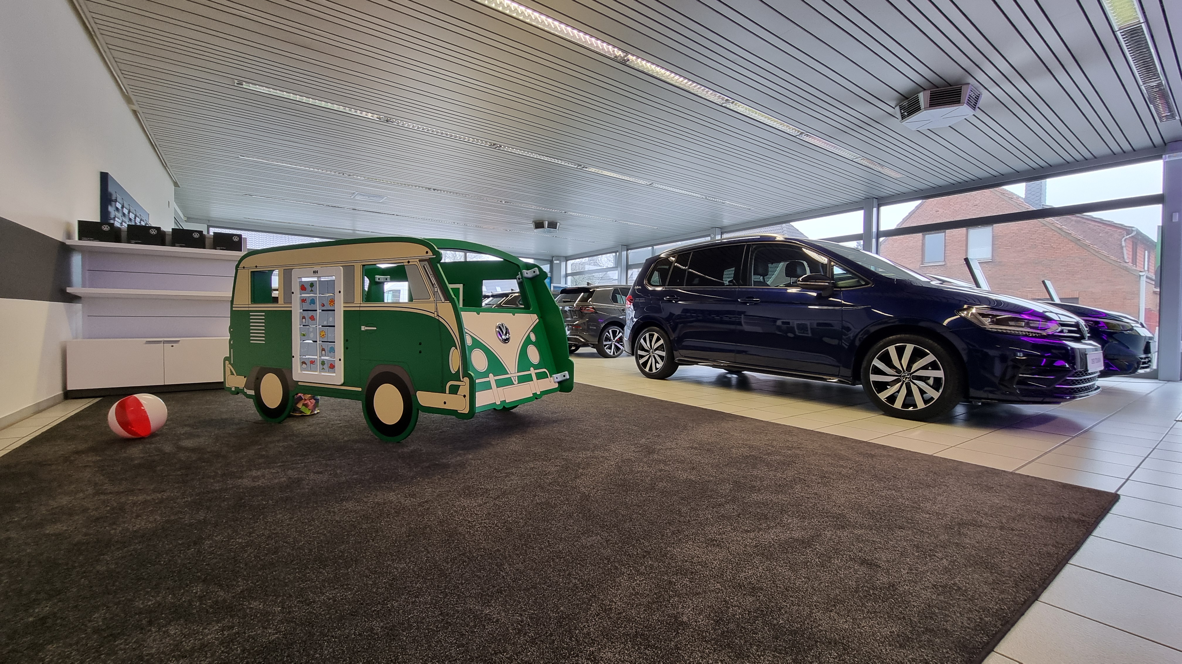 Groene IKC speelbus in de stijl van een klassieke Volkswagen, geplaatst op tapijt in een lichte showroom. Op de achtergrond staan diverse auto's opgesteld; links op de vloer liggen een strandbal en speelgoed.
