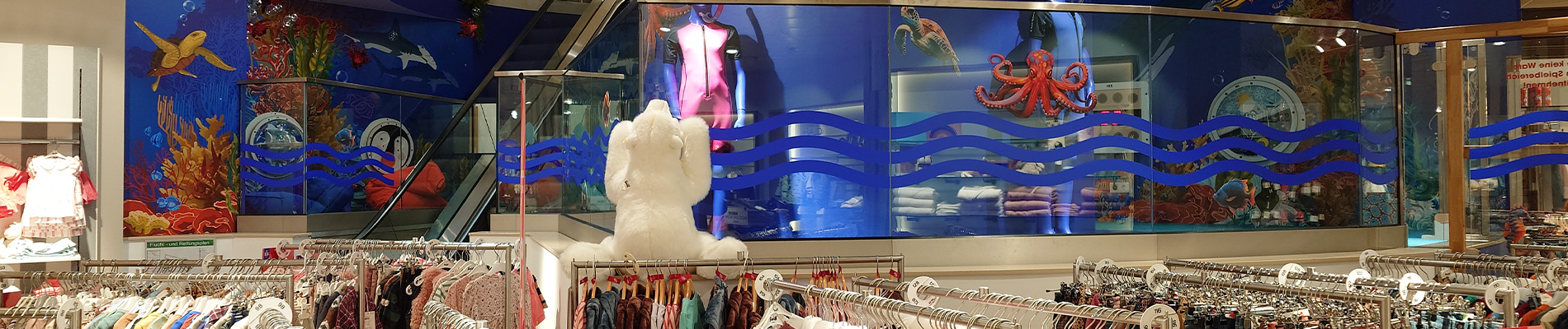 Kinderafdeling van Modehaus Baltz met kleurrijk onderwaterthema, inclusief muurschilderingen van een octopus, schildpad en vissen, naast de roltrap en kledingrekken